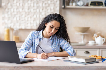 Young latin businesswoman writing at notepad while sitting in front of brand new laptop at kitchen interior