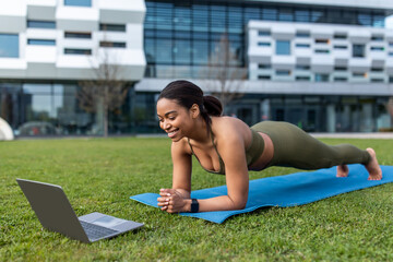 Sporty young black woman standing in plank, training to online sports video on laptop outdoors, copy space