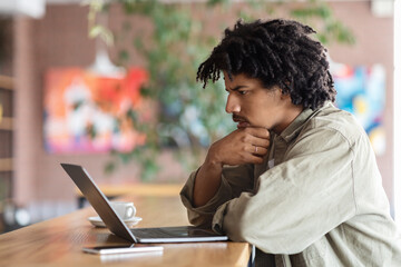 Pensive curly young black man looks at laptop screen at table in cafe interior