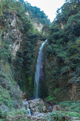A tall waterfall in Himalayas, along Annapurna Circuit Trek in Nepal. The water free falls from a high distance. The slopes of the mountain are overgrown with lush green plants.