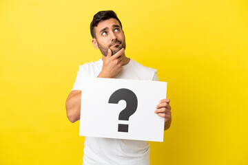 Young handsome caucasian man isolated on yellow background holding a placard with question mark...