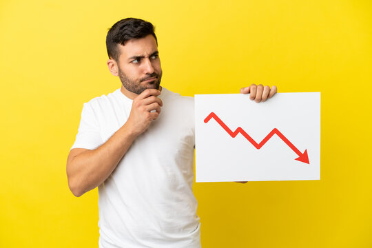 Young handsome caucasian man isolated on yellow background holding a sign with a decreasing statistics arrow symbol and thinking