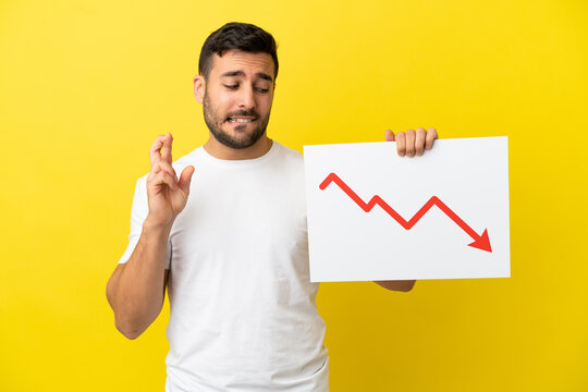 Young handsome caucasian man isolated on yellow background holding a sign with a decreasing statistics arrow symbol with fingers crossing