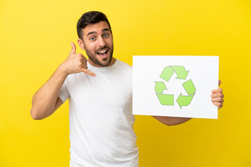Young handsome caucasian man isolated on yellow background holding a placard with recycle icon and...