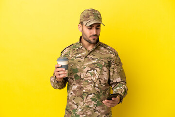Military man isolated on yellow background holding coffee to take away and a mobile