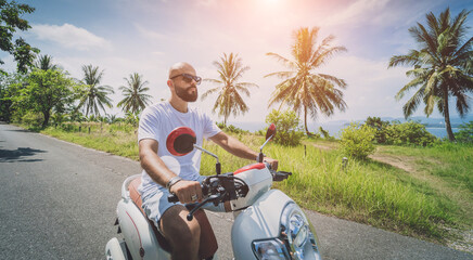 Stylish young man rides a motorbike on the road near the sea and palm trees