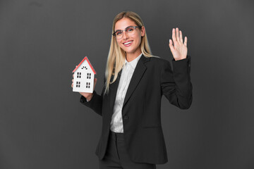 Blonde Uruguayan girl holding a house toy isolated on black background saluting with hand with happy expression