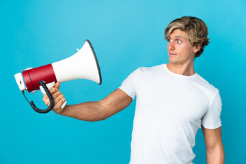 English man over isolated blue background holding a megaphone with stressed expression