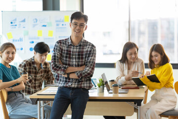 Portrait of happy smiling young businessman in office meeting room looking at camera while colleagues are meeting in the background.