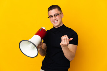Young caucasian man isolated on yellow background holding a megaphone and inviting to come with hand