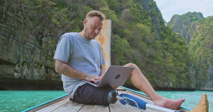 Disability freelancer caucasian man with artificial bionic legs prosthesis remote working anywhere on his laptop computer connected to office and relax by seeing beautiful view on summer vacation.