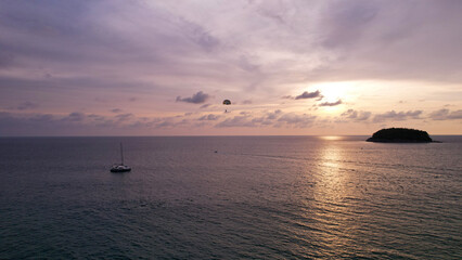 Parasailing at sunset with a view of the island. Parachute flight in the midst of a colorful sunset. Small waves, people relax on the beach, palm trees grow, there are hotels. Boats float. Asia Phuket