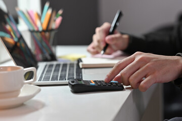 Close-up of male using a calculator and writing on a note next to a black screen laptop and a cup of coffee.Business and financial concepts.