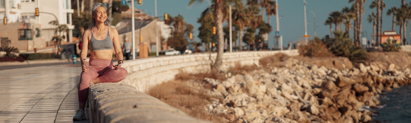 Strong active woman in sports clothes sitting by the seashore, holding buttle of water and looking ahead