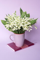 A bouquet of lilies of the valley stands in a mug on a pink notebook