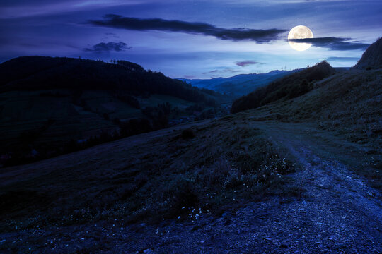 mountainous countryside landscape at night. rural fields on the hills. village in the distant valley. gorgeous sky with clouds glowing in full moon light