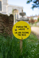 Pardon the weeds, we are feeding the bees sign placed in amongst wild flowers in a church yard