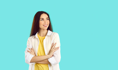 Happy attractive young brunette woman wearing casual tee and shirt standing with her arms crossed on turquoise studio background, smiling and looking at something on right copy space side