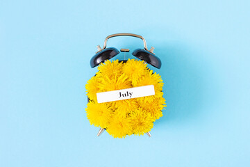 Calendar summer month July and alarm clock with yellow dandelions flowers on face, blue background. Top view Flat lay. Creative minimal concept July time. Top view, Flat lay, greeting card