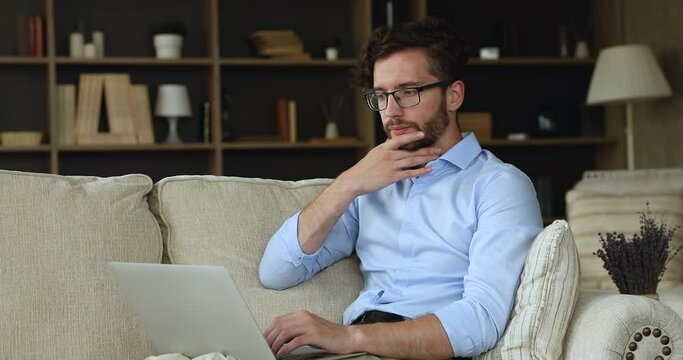 Millennial smiling man sit on sofa with wireless computer, texting message, thinks over creative online task looking pensive and thoughtful. Blogging, telecommute job using laptop, modern tech concept