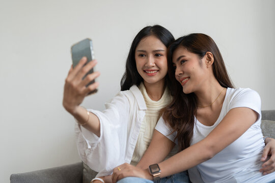 Asian beautiful lesbian or friends using mobile phone to take selfie together on couch. LGBT, Technology and Lifestyle Concept.