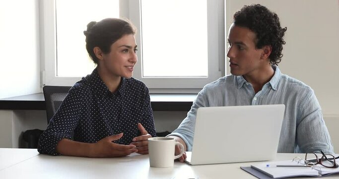 Young Latin male experienced worker mentor sit by laptop share professional skills with Indian female intern apprentice answer questions. Two diverse colleagues talk by pc overview electronic report