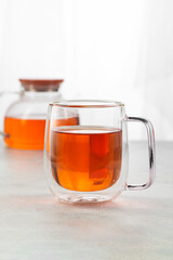 Glass cup of tea with teapot on light table against blurred background