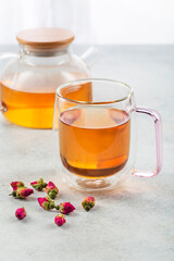 Glass cup of tea with teapot and rosebuds on light table against blurred background