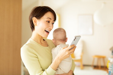 赤ちゃんを抱っこしながら携帯電話を操作する母親 - obrazy, fototapety, plakaty