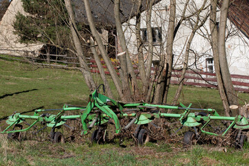 Farm equipment standing in a meadow. Farm buildings in the background.