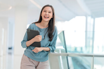 Fototapeta na wymiar asian smiling cheerful female digital nomad hand hold tablet device look at camera portrait shot,happiness smiling asia woman standing in office college corridor with positive smiling attitude