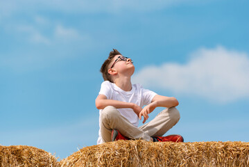 Naklejka na ściany i meble Happy teenage boy sits on haystack against blue sky. Child enjoys the fresh air and the sun in the village.