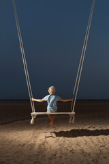 Portrait of boy on swing on the beach on dark sky background. Concept of childrens loneliness. Mystical back view portrait