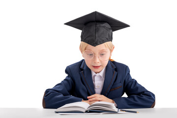 Young scholboy in school uniform and student hat sitting at table reading textbook. Education concept. Isolated on white background.