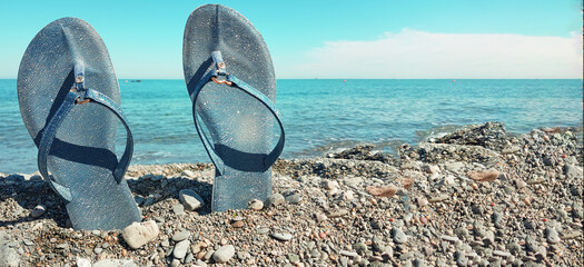 Blue women's beach shoes, standing in the pebbles against the background of the sea and the blue sky. Close-up, place for text, concept of a beach holiday by the sea, banner