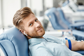 Portrait of smiling young man donating blood while lying in comfortable chair at med center and looking at camera - obrazy, fototapety, plakaty