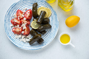 Plate with greek dolma or stuffed grape leaves, red tomatoes and feta, top view on a white stone background, horizontal shot