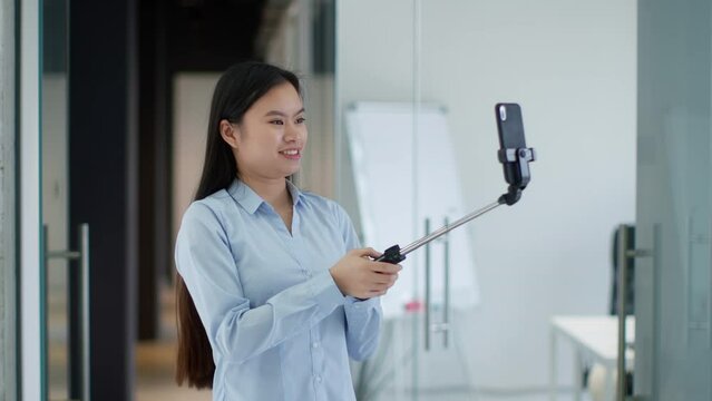 Young asian lady blogger recording video on smartphone, waving hand and talking to device, carrying cellphone in monopod
