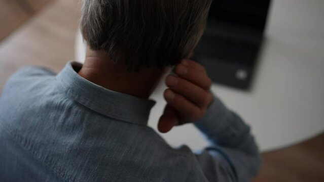 Close-up back view of unrecognizable mature gray-haired male experience severe pain in neck, rubbing it to relieve muscle tension, during working on laptop at home office. Shooting in slow motion.