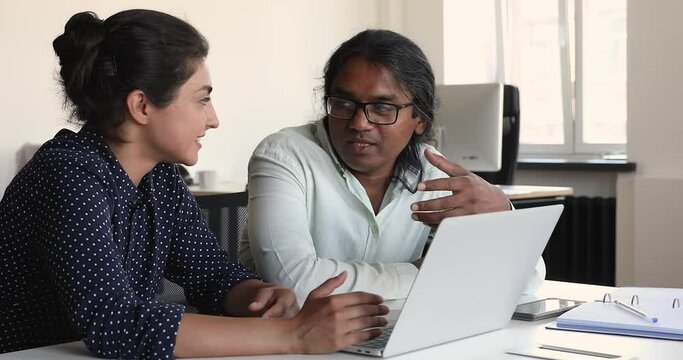 Two diverse colleagues of Indian ethnicity male teacher female student discuss task before laptop screen. Elder man supervisor project coordinator talk to younger female coworker review work result