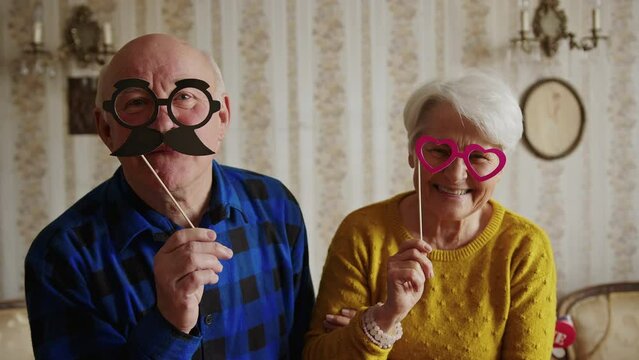 happy and cheerful senior couple holding fake heart-shaped glasses and mustache medium closeup indoor senior people support concept. High quality 4k footage