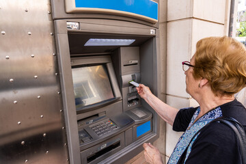 Elderly woman using an ATM in the street - obrazy, fototapety, plakaty