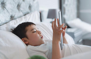 Happy Kid lying in bed holding tablet watching cartoon and chatting with friends on digital pad, Cute young boy playing games online on internet, Child relaxing in the morning before go to school