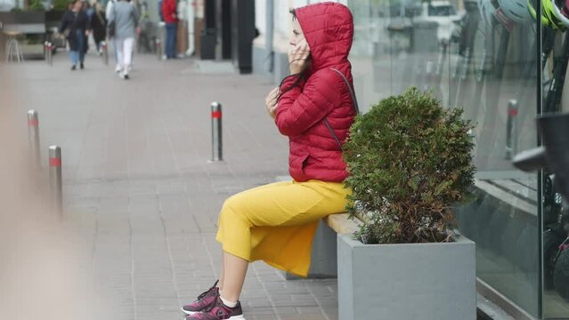 Young woman in a red jacket with a hood is talking by cell phone outdoors, sitting on street in city in windy day