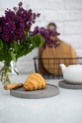 Fresh croissant on a grey plate on a table with a white teapot and lilacs