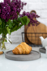 Fresh croissant on a grey plate on a table with a white teapot and lilacs