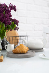 Fresh croissant on a grey plate on a table with a white teapot and lilacs