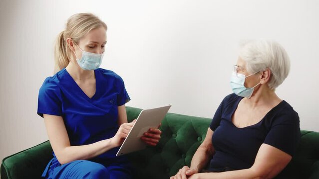 young health visitor with a mask holding a tablet and talking to her senior female patient medium shot indoor medical and healthcare concept. High quality 4k footage