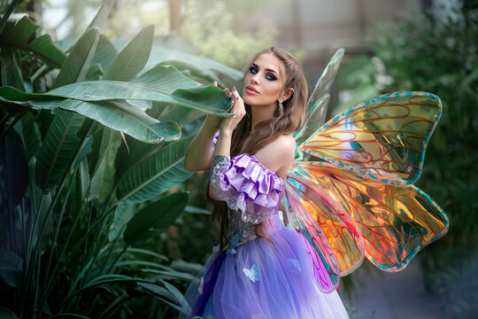 A beautiful woman is a little cute fairy with butterfly wings in a green exotic forest. Fantasy shooting of fairy-tale characters. Forest nymph, pixie girl in purple dress