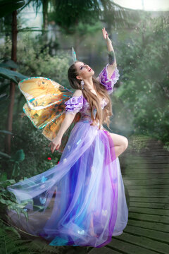 A beautiful woman is a little cute fairy with butterfly wings in a green exotic forest. Fantasy shooting of fairy-tale characters. Forest nymph, pixie girl in purple dress